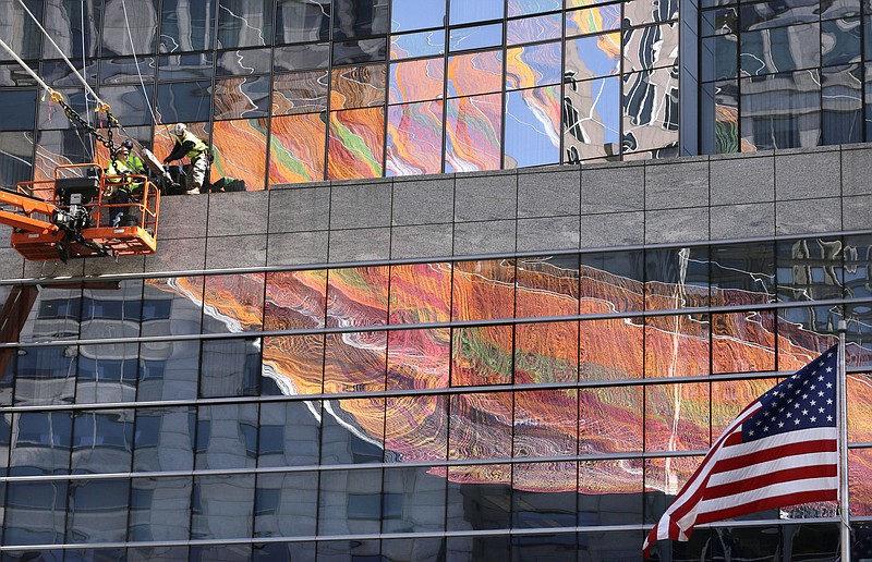 Workers, top left, secure lines that support a 600-foot sculpture, a portion of which is reflected in glass on a building, as it is suspended between high-rise buildings, Sunday, May 3, 2015, in Boston. The sculpture, that is to remain in place through October 2015, is made from over 100 miles of twine and utilizes over half a million knots in its construction.
