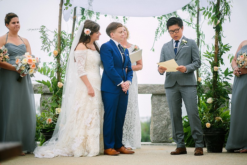 In this Aug. 22, 2015, photo, provided by Lisa Rigby shows Blake Keng, right, reading from Supreme Court Justice Anthony Kennedy's recent opinion on same-sex marriage during the wedding of Jillian Levine Smith, left, and Emily Smith, center, in Provincetown, Mass.