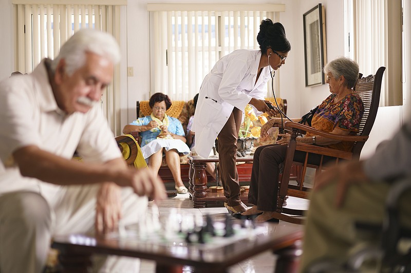Elderly people relax in a geriatric hospice while a doctor visits a patient, measuring her blood pressure.