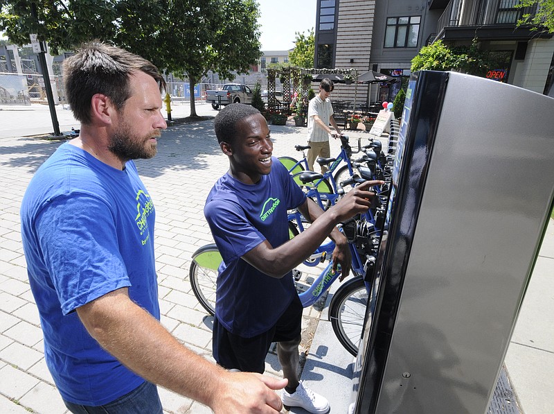 Lance MCrory, left, lead ambassador for Bike Chattanooga, instructs newly hired ambassador RaVontia Fuget on the operation of the kiosk located at the north end of the Walnut Street Bridge. Ben Taylor, background, of CDOT, returns a bike to its place. 