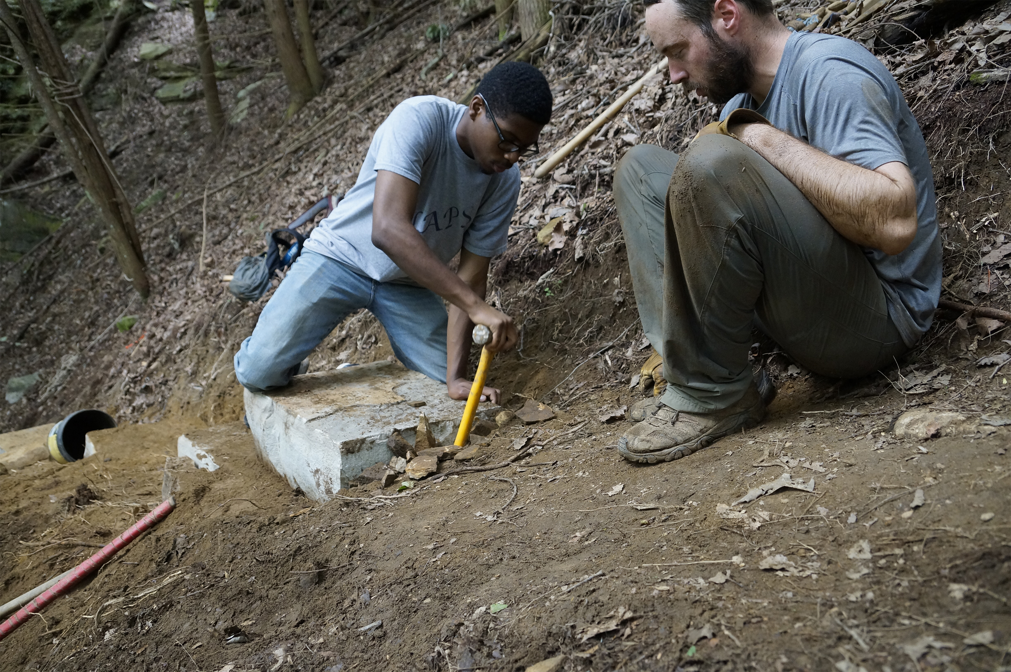 Staff writer Myron Madden, left, pounds gravel behind one of the massive staircase stones at McAlloyd Branch alongside Dustin Atkin, right. The gravel will prevent rainwater from displacing the structure.
