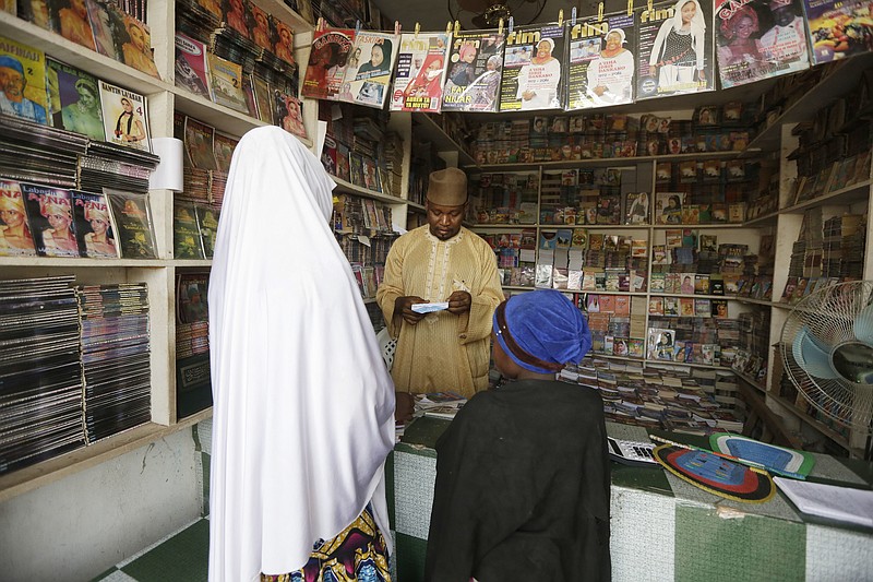 
              In this photo taken Sunday April, 3. 2016, Suleiman Maharazu, centre, the owner of Maharazu Bookshop, sells books to young girls in his shop in Kano, Nigeria. In the local market stalls are signs of a feminist revolution with piles of poorly printed books by women, as part of a flourishing literary movement centered in the ancient city of Kano, that advocate against conservative Muslim traditions such as child marriage and quick divorces.  dozens of young women are rebelling through romance novels, many hand-written in the Hausa language, and the romances now run into thousands of titles. (AP Photo/Sunday Alamba)
            