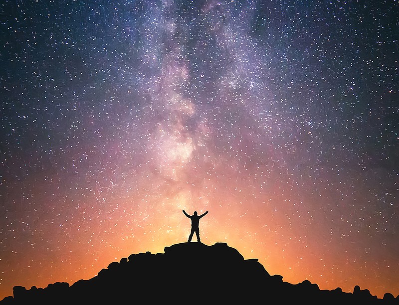 A person is standing on the top of the hill next to the Milky Way galaxy with his hands raised to the air.