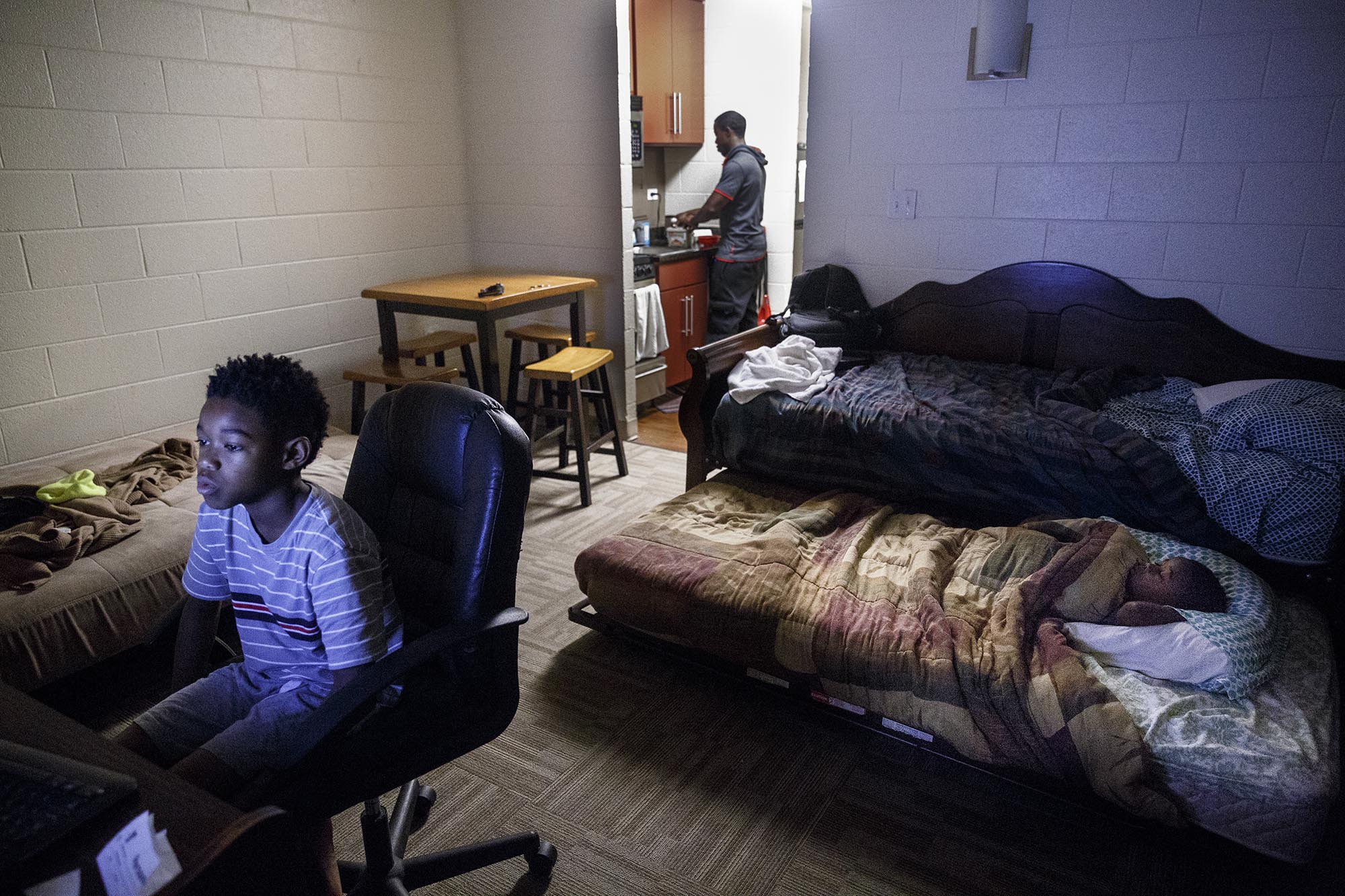 Tim makes oatmeal for his sons before school while Timothy, left, plays a reading game on the computer and Tariq sleeps in a pull-out trundle next to Tim's bed in his studio apartment.
