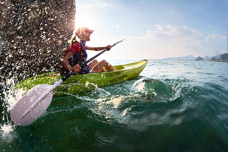 Young lady paddling hard the kayak with lots of splashes near the cliff at sunny day