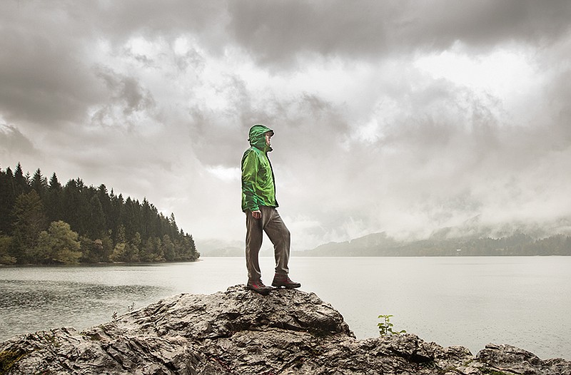 Man standing on a rock beside a dramatic mountain lake after a hike in the rainy, gloomy day. Active lifestyle, outdoor activities, moods and emotions concept.