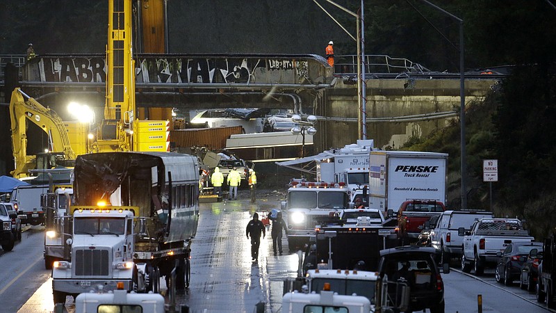 A damaged train car sits on a flatbed trailer at left as work continues to remove other cars at the scene of an Amtrak train crash onto Interstate 5 a day earlier Tuesday, Dec. 19, 2017, in DuPont, Wash. Federal investigators say they don't yet know why the train was traveling 50 mph over the speed limit when it derailed Monday, killing some people and injuring dozens. (AP Photo/Elaine Thompson)