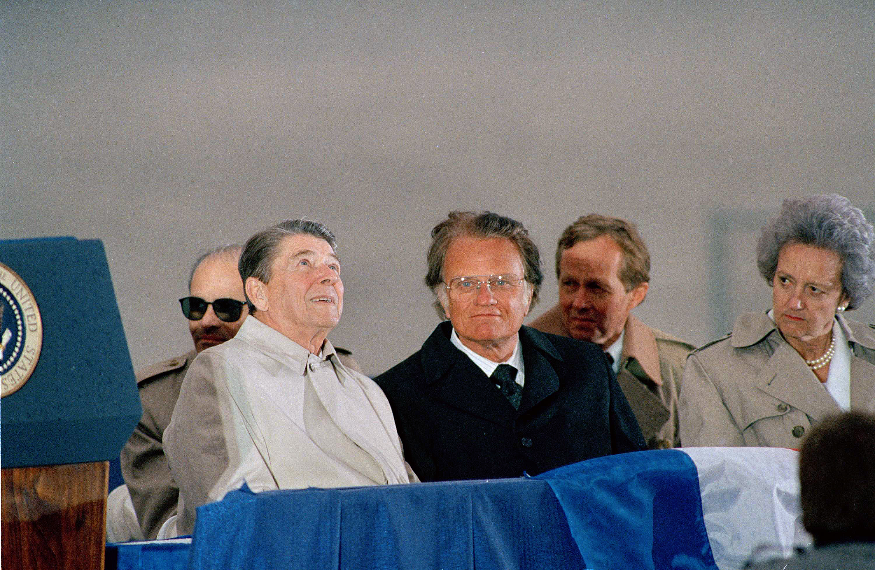 President Reagan, left, and Rev. Billy Graham, center, wait for a light rain to end at New York's Ellis Island, May 3, 1987, where Reagan delivered a speech during the opening ceremonies of the American Newspaper Publisher's Association annual convention. Others are unidentified. (AP Photo/Ron Frehm)
