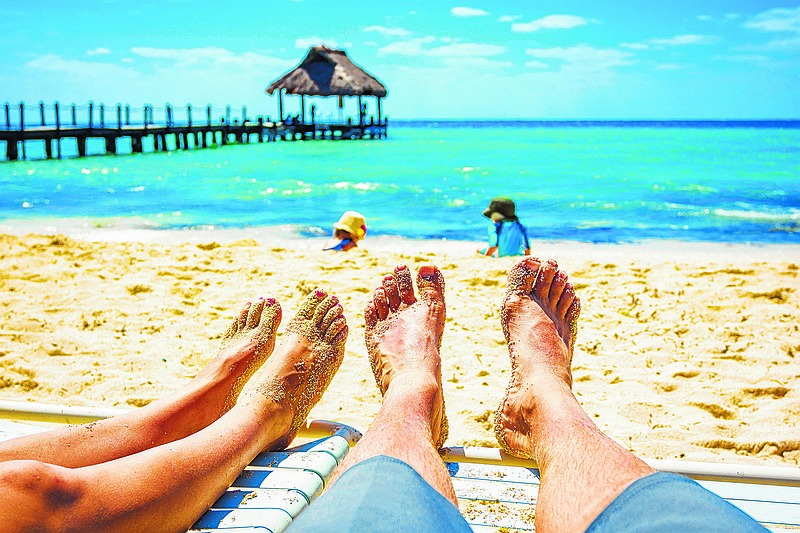 Sandy toes and feet of a couple on lounge chairs enjoying a beach vacation while watching their kids play in the sand. Tropical resort setting