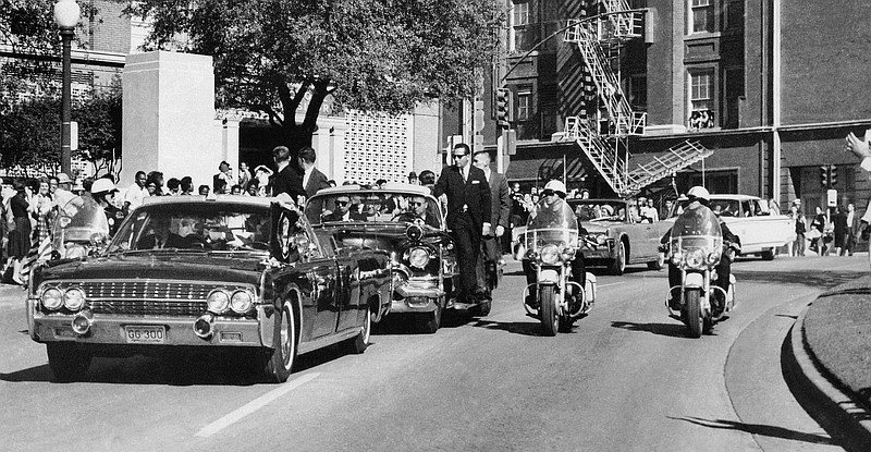 In this Friday, Nov. 22, 1963, photo, seen through the foreground convertible's windshield, President John F. Kennedy's hand reaches toward his head within seconds of being fatally shot as first lady Jacqueline Kennedy holds his forearm as the motorcade proceeds along Elm Street past the Texas School Book Depository in Dallas. Gov. John Connally was also shot.