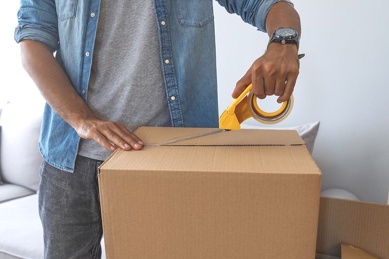 Close up of male hand packing cardboard box, concept moving house. House moving concept. Happy young couple moving into new apartment with packaging boxes. Male hands packing cardboard box with scotch moving out moving house tile / Getty Images