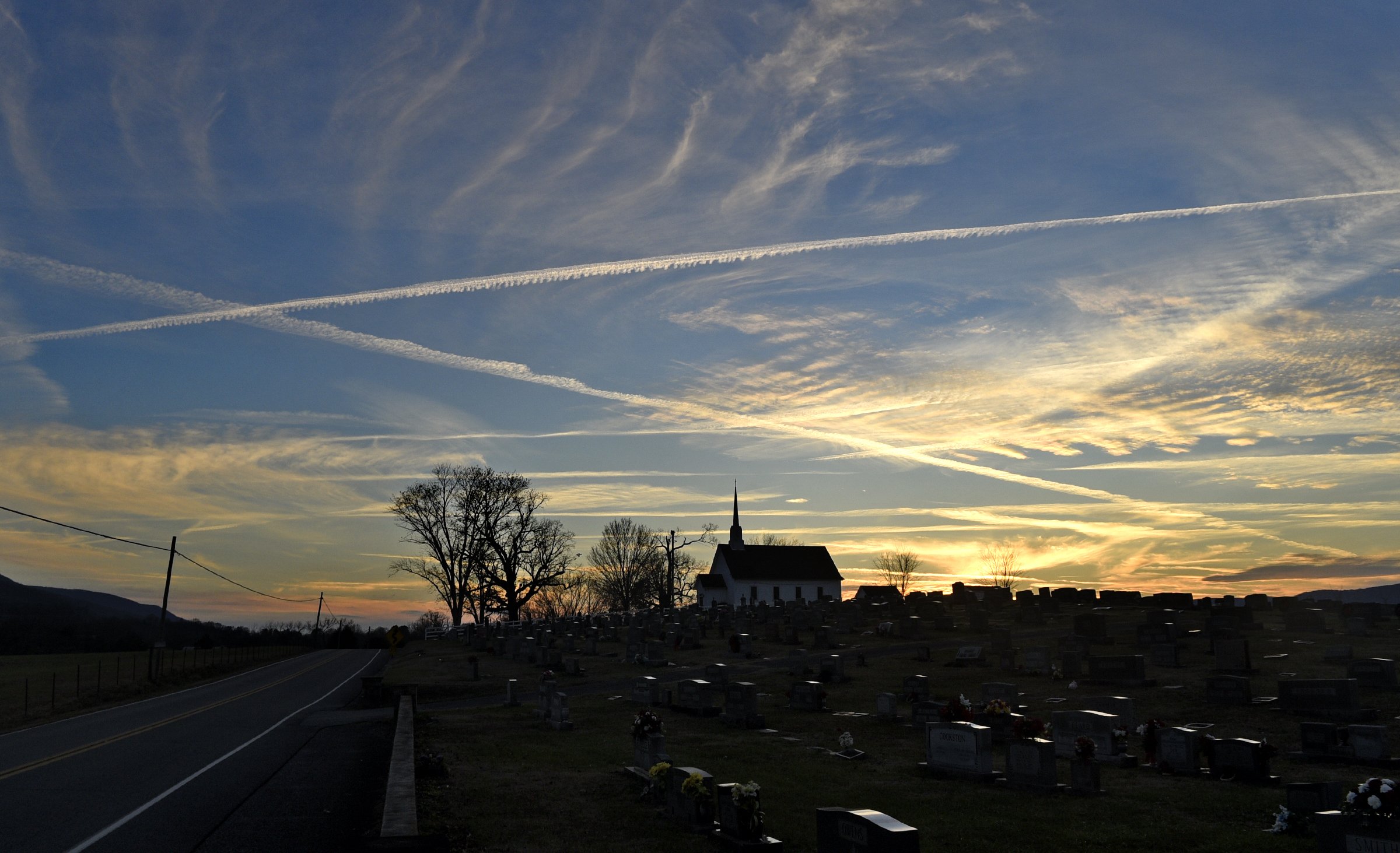 Clouds and contrails crisscross in the sky as the sun sets behind the Cumberland Plateau. Chapel Hill United Methodist Church stands in the foreground. The site has been a center for religious practice on the East Valley Road, east of Dunlap, in the Sequatchie Valley since 1851. The church can be seen along two cycling routes, the Daus Mountain Challenge and Middle Sequatchie Valley Loop, in a new database.