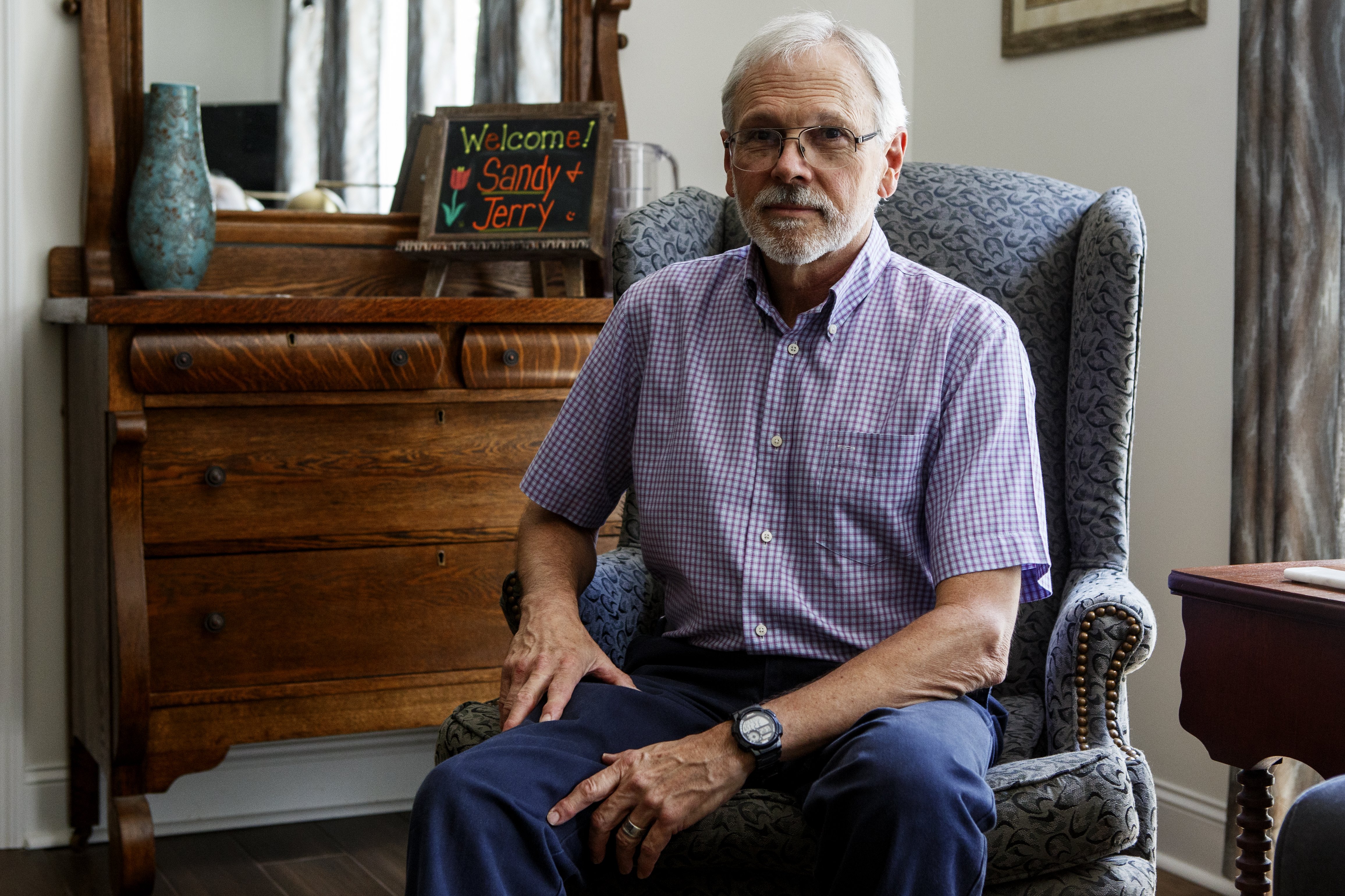 Owner Wallace Braud sits in the Monarch Room at St. Francis Cottage. The spacious rooms are furnished with multiple chairs and wooden side tables and cabinets.