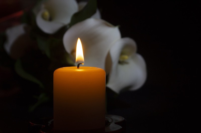 A close up of an orange candle and flame and lily flowers on a dark background. obit tile obituary death grave dead dies / Getty Images
