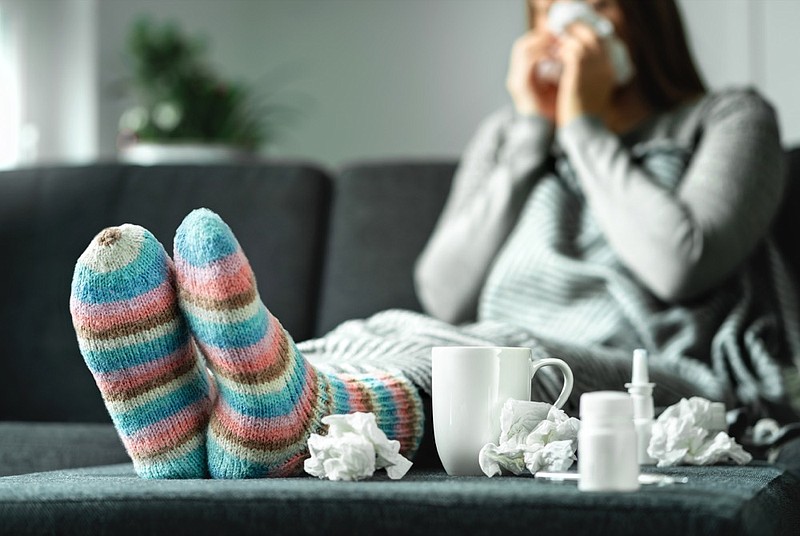 Sick woman with flu, cold, fever and cough sitting on couch at home. Ill person blowing nose and sneezing with tissue and handkerchief. Woolen socks and medicine. Infection in winter. Resting on sofa. flu tile sickness health ill illness cold tile / Getty Images
