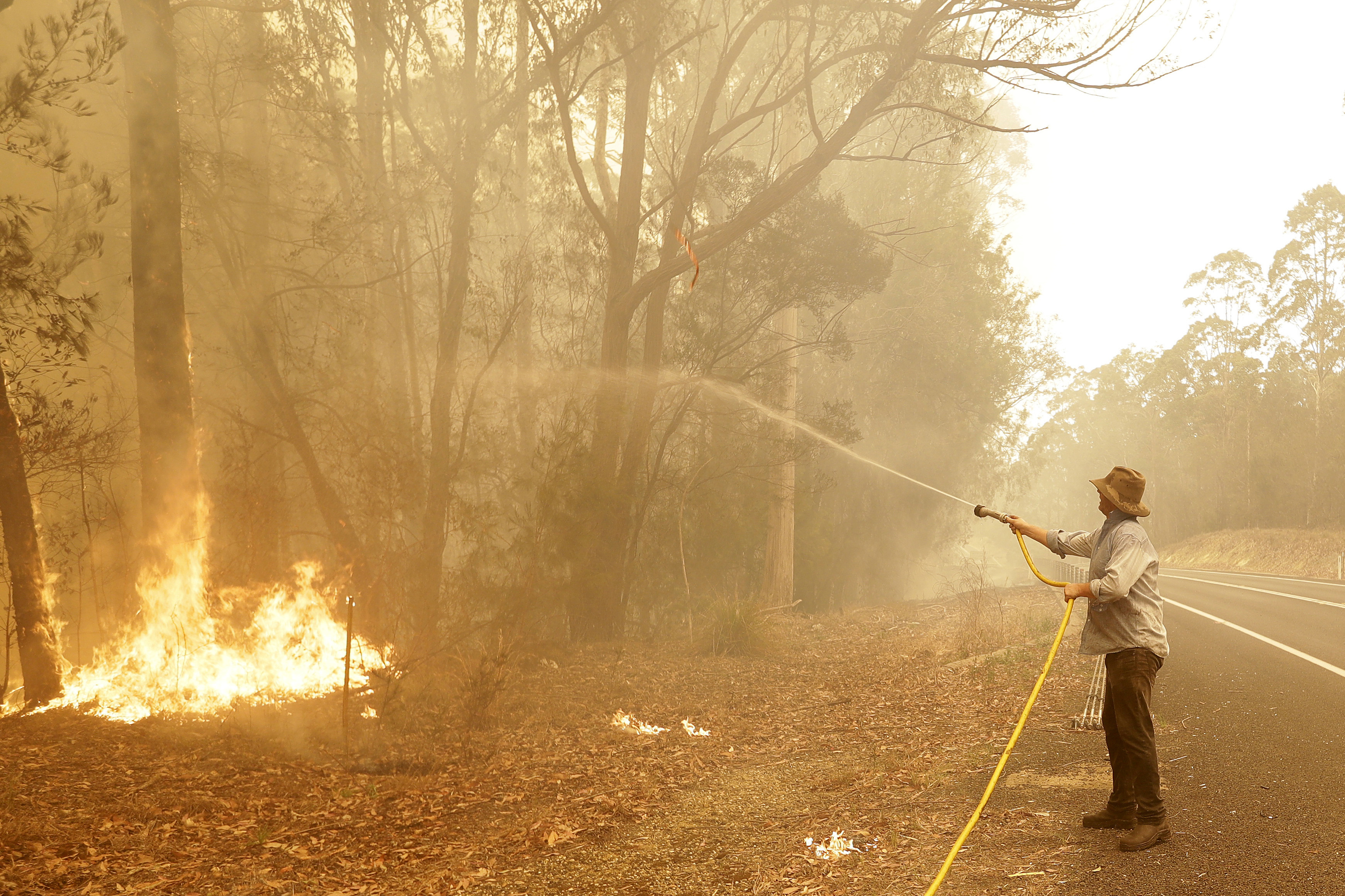 A man uses a water hose to battle a fire near Moruya, Australia, Saturday, Jan. 4, 2020. Australia's Prime Minister Scott Morrison called up about 3,000 reservists as the threat of wildfires escalated Saturday in at least three states with two more deaths, and strong winds and high temperatures were forecast to bring flames to populated areas including the suburbs of Sydney. (AP Photo/Rick Rycroft)