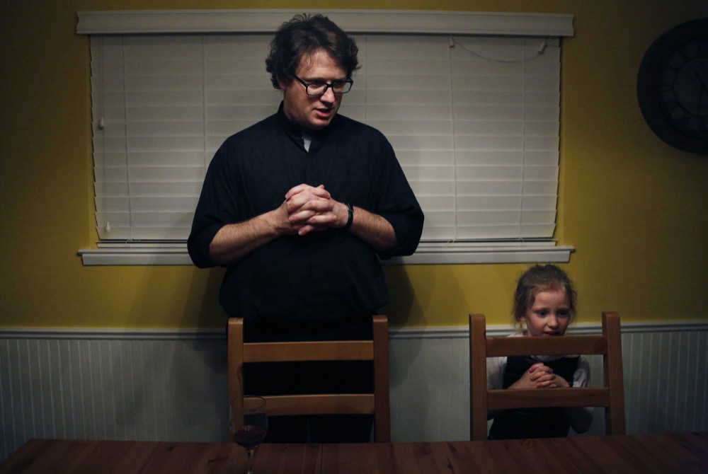 In this Feb. 7, 2020, photo, converted Catholic Priest Joshua Whitfield, left, his daughter Zoe-Catherine, 5, and the rest of his family stand behind their chairs to say grace before dinner in north Dallas. (AP Photo/Jessie Wardarski)