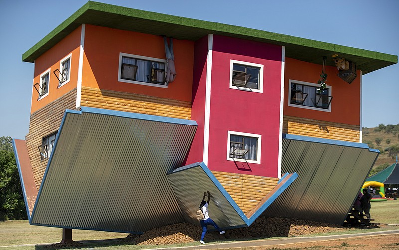 In this photo taken Friday, March 5, 2020 a visitor pretends to hold the structure up at the upside-down house in Hartbeespoort, South Africa. With its roof on the ground and it floor in the air, the upside down house is attracting tourists who want to see the world from a different perspective. (AP Photo/Themba Hadebe)