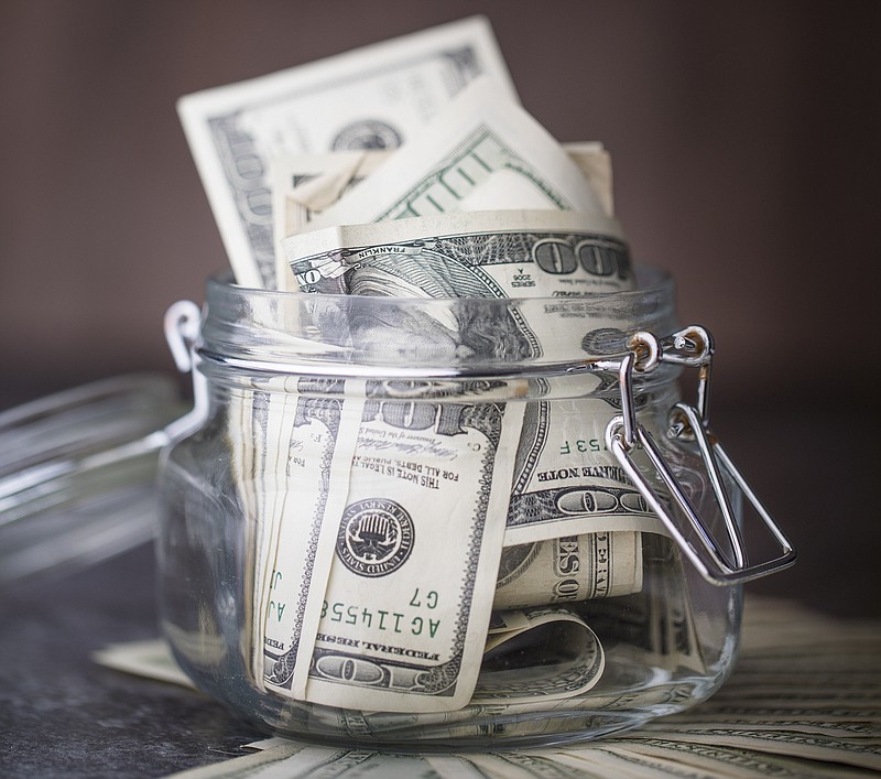 US 100 dollar bills in a glass jar, with more bills on the table under the jar. With a brown wooden background. saving tile money tile emergency saving / Getty Images
