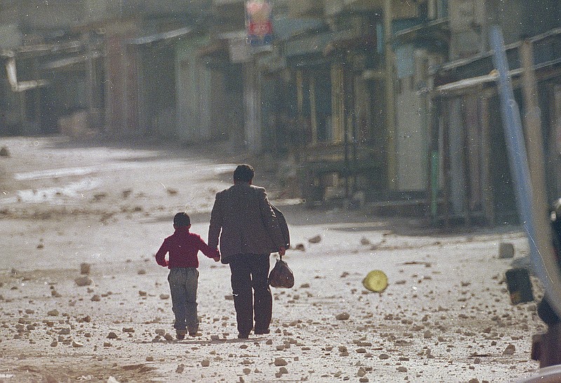 FILE - In this Dec. 11, 1987 file photo, a man holds his son's hand as they make their way along a debris littered street in the Balata refugee camp near Nablus in the occupied West Bank after a curfew was imposed on the camp. Western countries are reeling from the coronavirus pandemic, awakening to a new reality of economic collapse, overwhelmed hospitals and home confinement. But for millions across the Middle East and in conflict zones elsewhere, much of this is familiar. (AP Photo/Max Nash, File)

