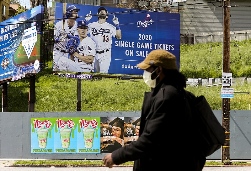 AP photo by Damian Dovarganes / A pedestrian wears a hat and a face mask on Sunset Boulevard in the Echo Park neighborhood of Los Angeles, with a billboard in the background advertising tickets for a Dodgers season that has yet to start and might not due to the COVID-19 pandemic.