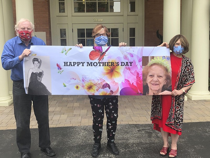 This May 3, 2020, photo released by Shelly Solomon shows, from left, Steve Turner and his sisters, Carla Paull and Lisa Fishman, holding up a Mother's Day banner emblazoned with images of their mom, Beverly Turner, in front of her assisted living facility in Ladue, Missouri. They were "practicing" how their Mother's Day surprise will look on the holiday as their mother peers down from a window. Isolation due to the coronavirus outbreak has led mothers and offspring to find creative ways to celebrate. (Shelly Solomon via AP)