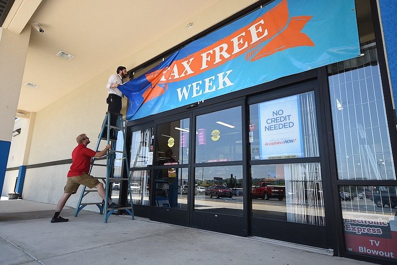 In this 2019 staff file photo, Mark Johns, left, steadies a ladder for Electronic Express assistant manager Nathan Adams to re-secure the "tax free week" sign that blew loose from the original hooks at the Hixson retailer. "We've had this sign up all week," Adams said. / Staff file photo by Tim Barber