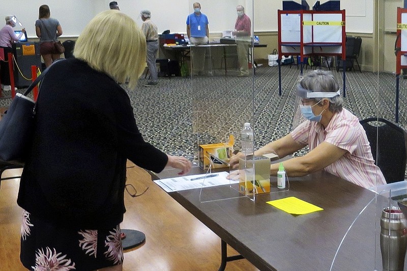 FILE - In this July 14, 2020, file photo, election worker Adonlie DeRoche, seated, wears a mask and face shield behind Plexiglas for safety during the coronavirus pandemic, while handing a ballot and single-use pen to a voter during the primary election in Portland, Maine. Thousands of U.S. election officials are busy sharing creative ideas they hope will keep voters and polling places safe from infection. (AP Photo/David Sharp, File)