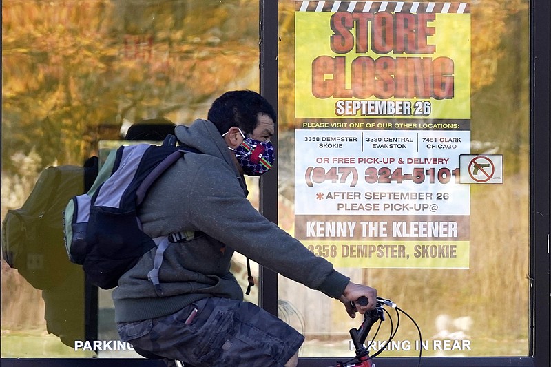 In this Friday, Nov. 6, 2020, file photo, a man wearing a face mask rides a bicycle past a closed store in Wilmette, Ill. States in the U.S. are renewing their push for more federal money to deal with the fallout from the coronavirus outbreak and to help them distribute a vaccine when one becomes widely available sometime in 2021. (AP Photo/Nam Y. Huh, File)