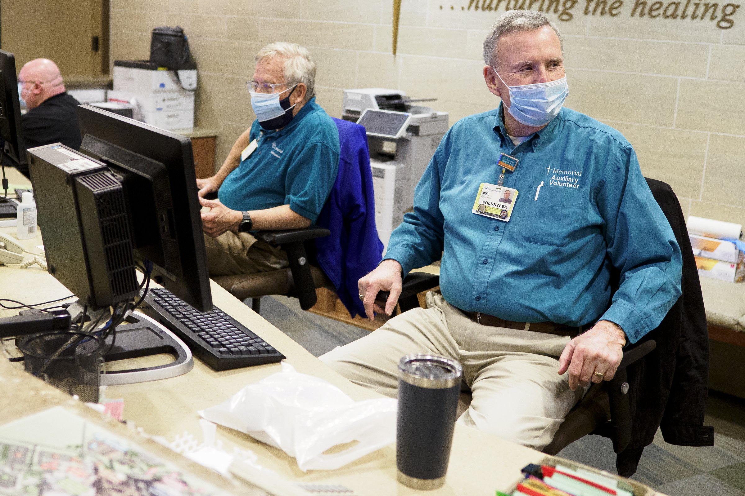 Staff photo by C.B. Schmelter / Volunteers John Vass, center, and Mike Doubleday sit behind the desk waiting to help at CHI Memorial Hospital on Monday, Feb. 1, 2021 in Chattanooga, Tenn. The hospital welcomed back volunteers who have been fully vaccinated.