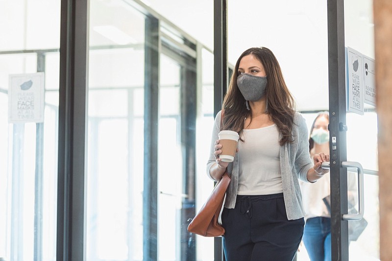A mid adult businesswoman enters her office during the COVID-19 pandemic. A colleague is walking behind her, practicing the proper social distancing protocol. The businesswomen are wearing protective face masks. work office covid tile / Getty Images
 